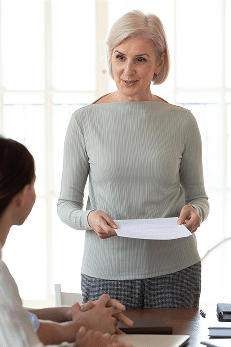 Older woman stands in a meeting room holding papers and speaking