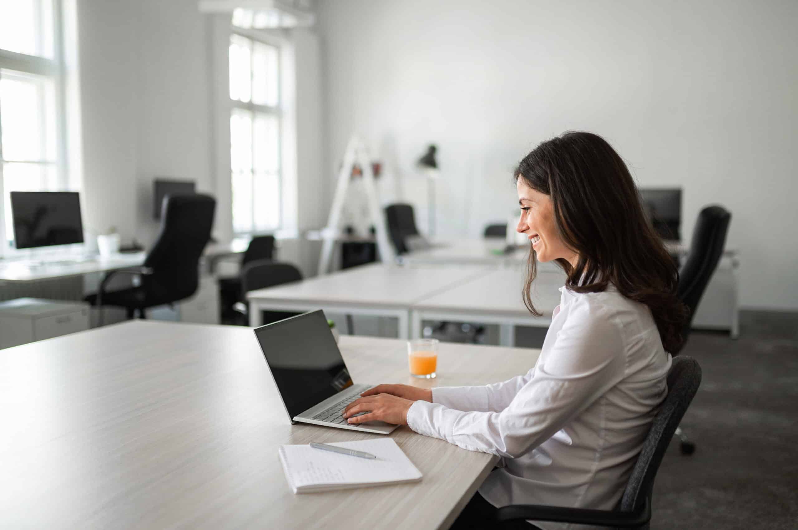 Woman smiling while working on a laptop in a bright office